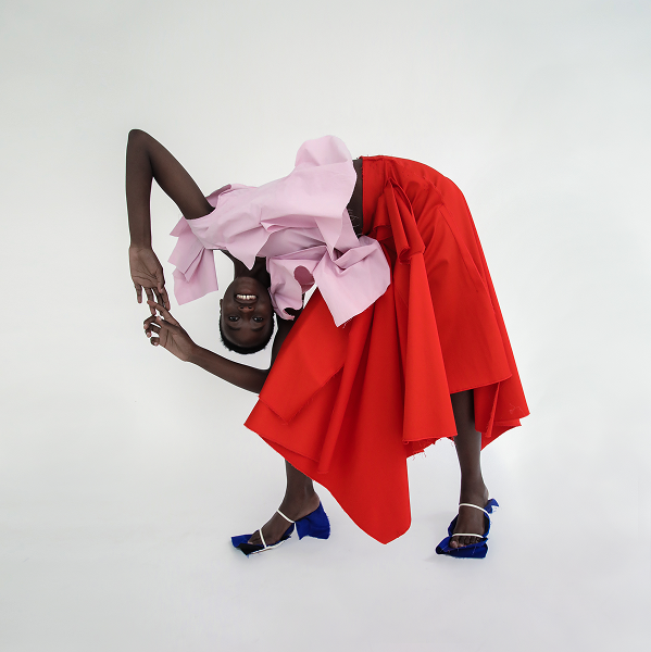 Editorial portrait — model folded forward in a draped red and pink ensemble with cobalt heels