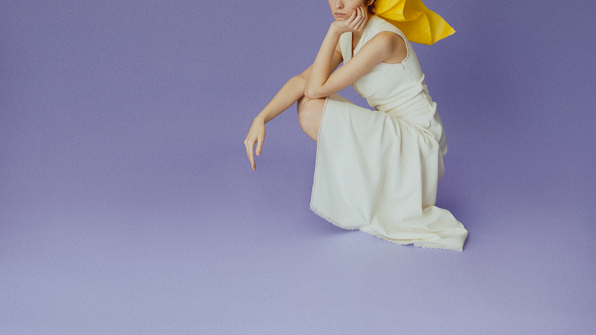 Model seated in a flowing ivory dress against a soft lavender backdrop
