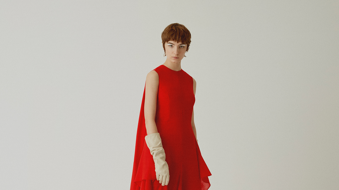 Model wearing the signature red pleated dress against a warm off-white studio backdrop